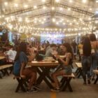 Pessoas sentadas em bancos com mesas, comem e aproveitam shows em rua fechada e iluminada