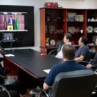 Prefeito e autoridades da Suíça e de Joinville em volta de mesa, no gabinete, olhando uma televisão