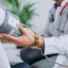 Profissional da saúde com jaleco e estetoscópio, afere pressão de uma pessoa por meio de um aparelho no braço do paciente