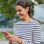 Mulher com cabelo amarrado e blusa listrada segura celular e sorri ao olhar o aparelho