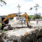 Local da obra, com retroescavadeira e homens com capacete e uniforme trabalhando na rua