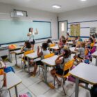 Professora na frente de quadro em sala de aula explica conteúdo para alunos sentados em cadeiras na frente de carteiras