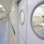 Mulher profissional da saúde com máscara, luvas e uniforme caminha em corredor de hospital