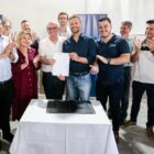 Prefeito e padre seguram papel, posam para foto, ao lado de autoridades