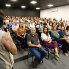Mulher com colete do Ministério da Saúde fala no microfone em pé voltada para plateia