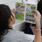 Menina de costas, com rabo de cavalo no cabelo, lê gibi com cotovelos apoiados em mesa