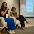 Duas mulheres sentadas em poltronas em um palco, uma delas segura um microfone e fala com a plateia