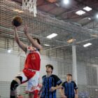 Atleta pula com bola de basquete para arremessar na cesta enquanto dois atletas adversários observam