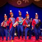 Grupo de músicos vestidos de soldados com figurinos azul e vermelho posam para foto em palco e sorriem