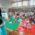 Mulher segura livro e conta história para crianças sentadas no chão da biblioteca