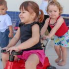 Menina brinca em motoca empurrada por outra menina pequena e outras crianças observam e brincam ao redor