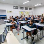 Agente da Defesa Civil, de colete laranja, fala para alunos em sala de aula