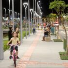 Criança anda de bicicleta em passeio público, pessoas caminham e conversam sentadas em bancos