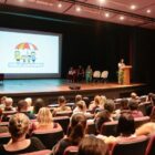 Mulher fala em púlpito em palco e plateia assiste sentada em poltronas