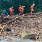 Cinco homens da Defesa Civil, com uniforme laranja, andam em área atingida pela chuva