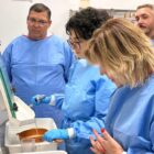 Quatro pessoas com avental azul observam uma delas manusear equipamento de laboratório