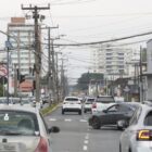 Rua com quatro pistas, um canteiro no meio, postes com fios e carros trafegando
