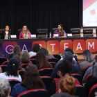 Um homem em pé fala ao microfone em palco, três mulheres estão sentadas. Na frente, há uma plateia sentada.