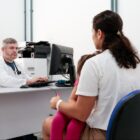 Homem sentado de jaleco branco e estetoscópio em volta do pescoço em frente a um computador olha para mulher com criança no colo em consulta médica