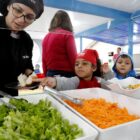 Três crianças de Centro de Educação Infantil em frente a pratos com saladas ao lado de professora com touca no cabelo e uniforme de chef de cozinha
