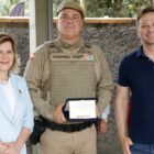 Policial militar fardado segura placa e posa para foto ao lado do prefeito e da vice prefeita