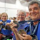 Dois homens e uma mulher com camisetas azuis sorriem e mostram medalhas penduradas no pescoço
