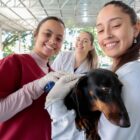 Duas mulheres seguram um cachorro e sorriem para foto, uma outra mulher atrás também olha para a câmera e sorri