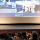 Mulher loira veste calça jeans e blusa preta segura microfone em palco e fala para plateia sentada