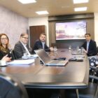 Oito pessoas, entre homens e mulheres, sentados ao redor de uma mesa conversam, há um telão atrás da mesa