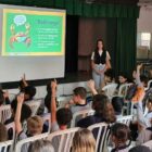 Um homem e uma mulher em pé ao lado de um telão fazem palestra para alunos sentados em cadeiras na frente deles