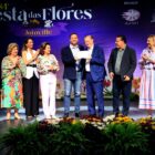 Oito pessoas, entre homens e mulheres, em palco. Prefeito e homenageado seguram certificado