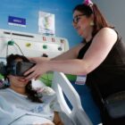 Paciente em cama de hospital usa óculos de realidade virtual segurado por uma profissional de saúde