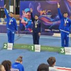 Três atletas com uniforme azuis com medalhas e braços levantados