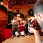 Papai Noel com duas crianças ao lado sentados numa poltrona vermelha, ao lado de árvore de Natal. Homem tira foto