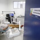 Duas profissionais com roupa branca, touca e máscara atendem paciente deitado em cadeira em consultório odontológico