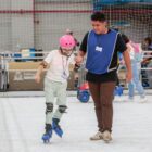 Menina com capacete rosa e patins anda em pista de patinação no gelo de mãos dadas com um instrutor. Há outras pessoas e crianças patinando também