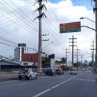 Rua com carros trafegando em duas pistas, comércios e casos ao redor, fiação elétrica e placas
