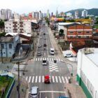 Foto aérea de uma rua com carros trafegando em duas pistas, além de casas e comércios