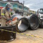 Homem com uniforme e equipamentos de segurança trabalha com equipamento perto de tubulações e caminhão em rua
