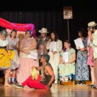 Grupo com dez pessoas em palco encenam peça de teatro com figurinos e cenário