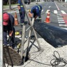 Três homens uniformizados trabalham em rua em bueiro com equipamentos