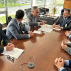 Sete homens, todos de terno e gravata, sentados em volta de uma mesa conversam