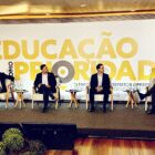 Quatro homens com terno sentados em cadeiras em palco, um deles segura um microfone e fala para a plateia