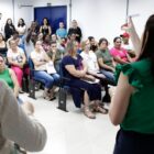 Duas mulheres de costas, em pé, olham para pessoas sentadas em cadeiras. Uma das mulheres segura um papel no alto e mostra para as pessoas
