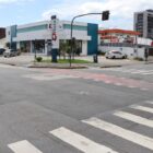 Cruzamento de duas ruas, com faixas de pedestre no chão, semáforos, carro trafegando. Em uma das esquinas, há uma farmácia