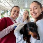Duas mulheres jovens seguram um cachorro preto com luvas e uma terceira está atrás delas, todas olham para a câmera e sorriem