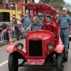 Carro vermelho antigo do Corpo de Bombeiros com homens desfila em avenida