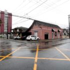 Rua com sinalização viária em amarelo no chão, em uma das esquinas uma casa com pintura vinho e carros estacionados ao lado