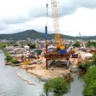 Imagem aérea de terreno onde está sendo construída ponte, com grandes equipamentos e guindaste