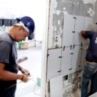 Dois operários com capacetes trabalham em obra em escola, eles instalam azulejos